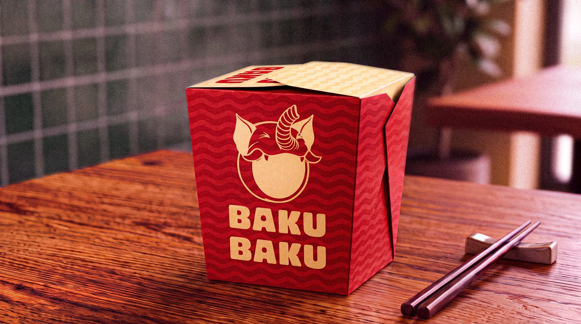A red Baku Baku noodle box on a wooden table, the mark and wordmark in cream over the noodle wave, chopsticks beside it