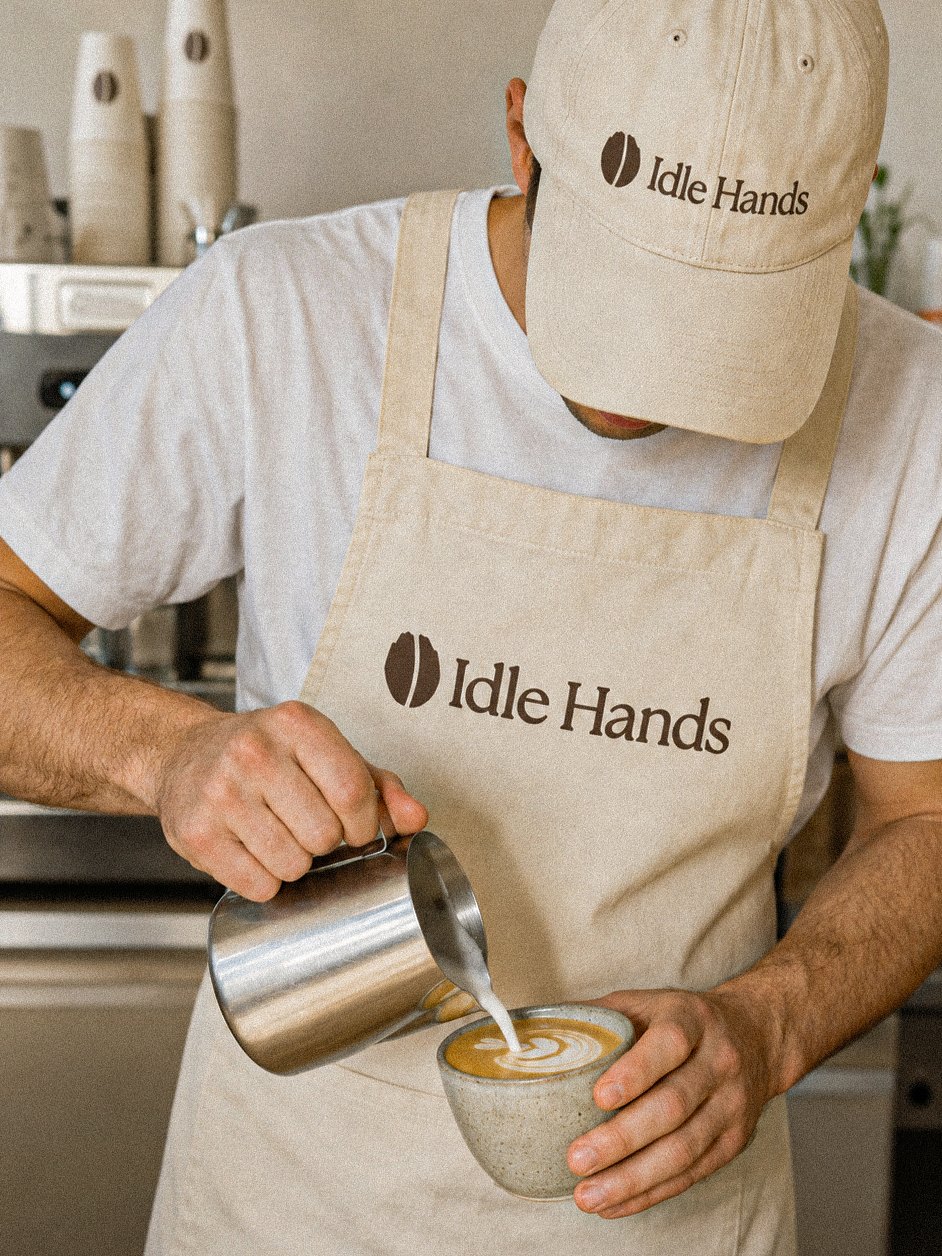 A barista in the Idle Hands apron and cap pouring latte art into a stoneware cup