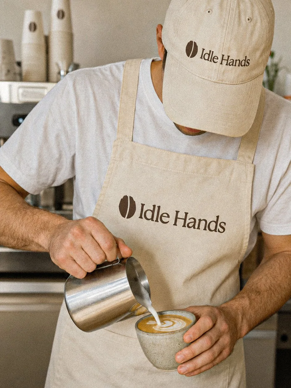A barista in the Idle Hands apron and cap pouring latte art into a stoneware cup